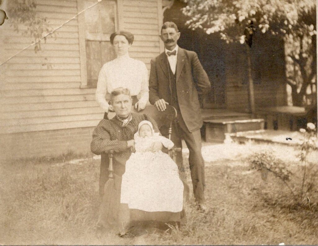 Sepia-toned photo of two adults standing, one sitting holding a baby.