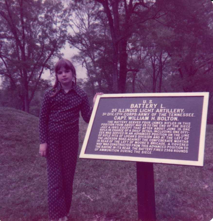A young white girl in a 1970s pantsuit stands next to a plaque describing the actions of the 29th Illinois Light Infantry at the battle of Vicksburg