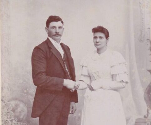 An old photo of a white couple holding hands, a man wearing a dark suit, a woman wearing a white dress.